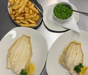Grilled fish with lemon and herb garnish, bowl of peas and freshly cooked chips.