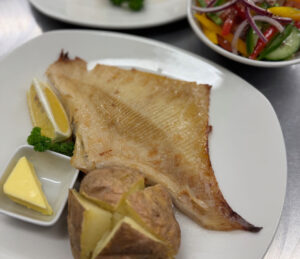 Grilled fish with a jacket potatoes, butter and lemon garnish. In the corner is a freshly prepared salad.
